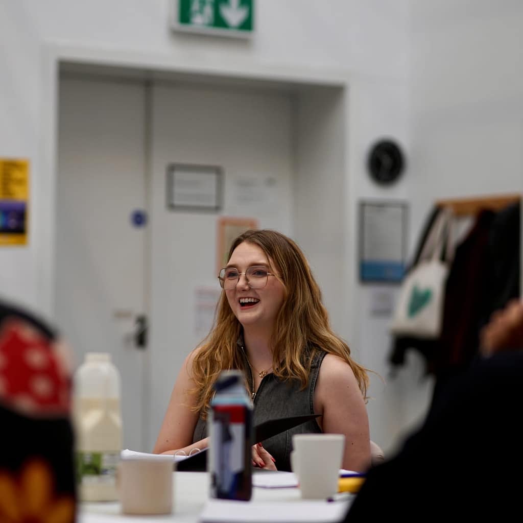 The artist has long brown hair and classes, talks at a table whilst smiling