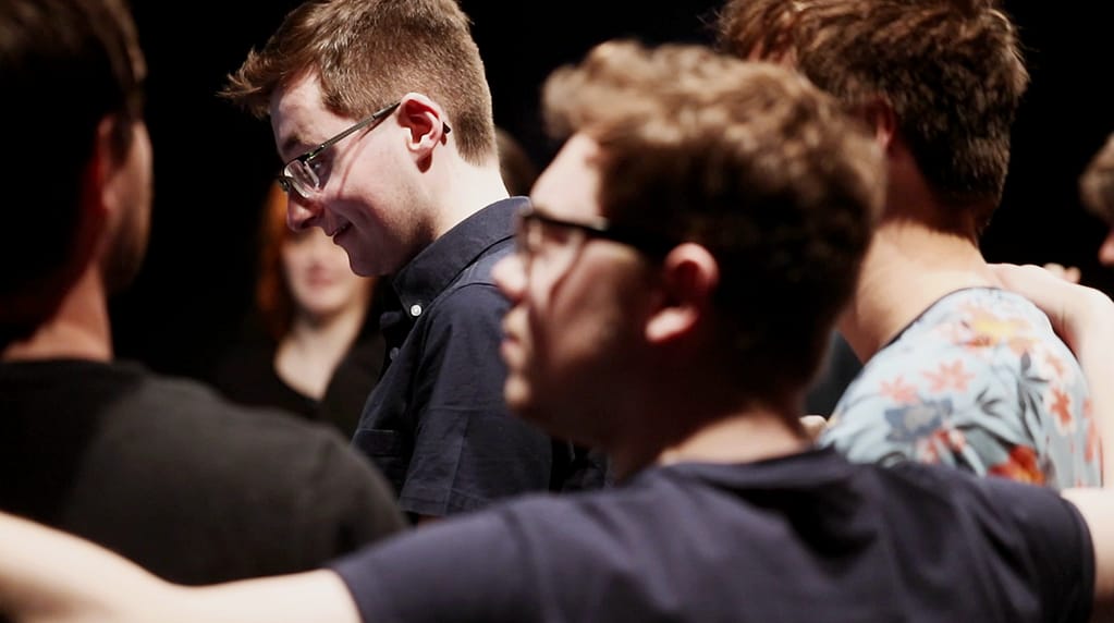 In focus, a young person with short hair, glasses and a navy blue shirt stands with a side profile looking downwards. Out of focus, in the foreground, are the back of some other young people in different coloured t-shirts with their arms round one another. A girl with ginger hair is out of focus in the background