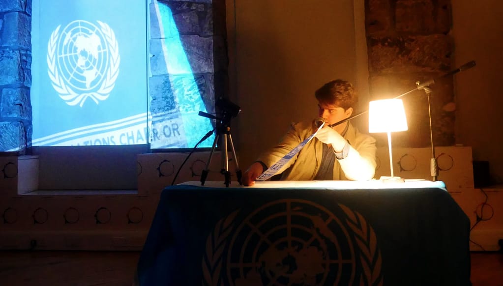 A white male with short dark hair sits at a desk with his elbows leaning on the desk behind a bright, illuminating lamp. He speaks into a microphone in front of him while holding up a poster to the left of him.