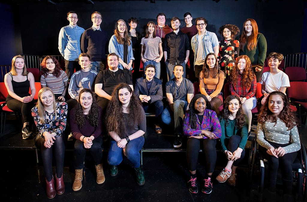 A cast of performers sit and stand against banked seating in a black box theatre with bright light reflecting off their faces