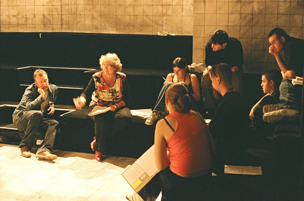 A group of people sit on varying heights of banked stage seating. They hold scripts in their hands and look intently at one another. One person has their knees up to their chest and is writing