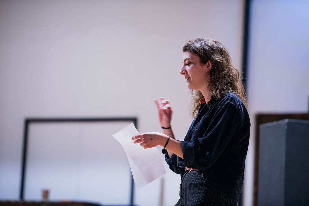 A young person dressed in a black shirt and trousers with long brown hair holds a sheet of paper in one hand and rehearses lines, gesturing with their other hand