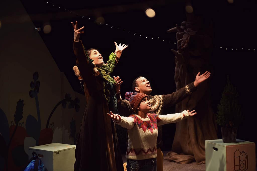 Three young performers look towards the sky with their arms outstretched, with dazzled/amazed looks on their faces. They wear different coloured knitted jumpers. There is a prop tree in the background, and wooden boxes surround them on the floor.