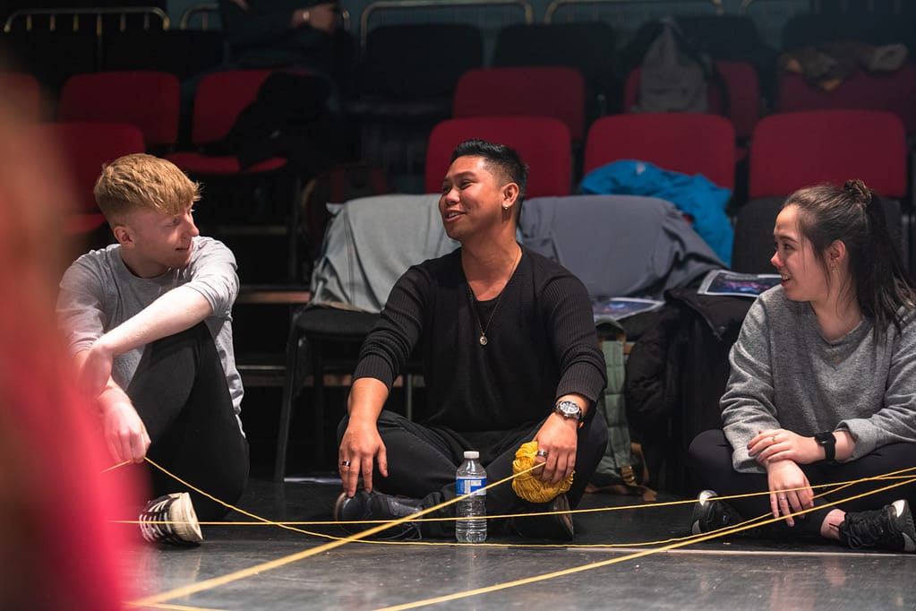 A young man with short dark hair sits cross-legged, wearing all black and holding a ball of yellow yarn in his hand. Two young people sit either side of him watching him as he talks.