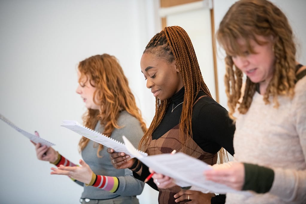 3 woman stand in a row looking down at scripts they are holding in their hands. 2 are out of focus to the left and right of the frame. The black woman in the centre of the frame smiles with one hand on her hip. She wears a black turtleneck and brown dungarees.