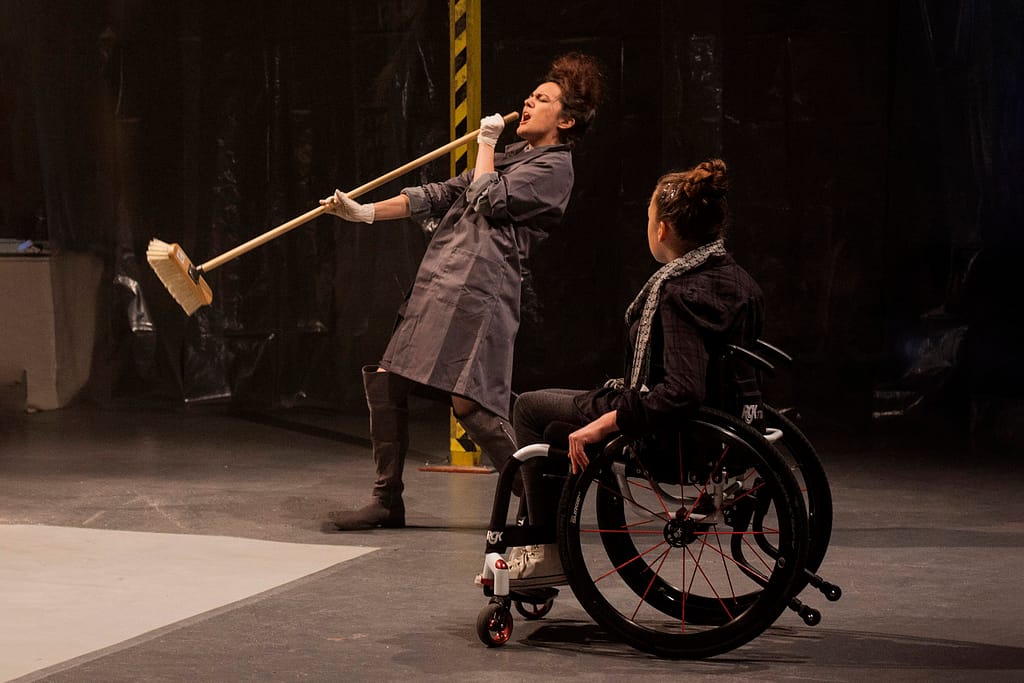 A performer in a wheelchair has their back to the camera and watches a performer dressed in overalls in the background mime singing into a broom passionately