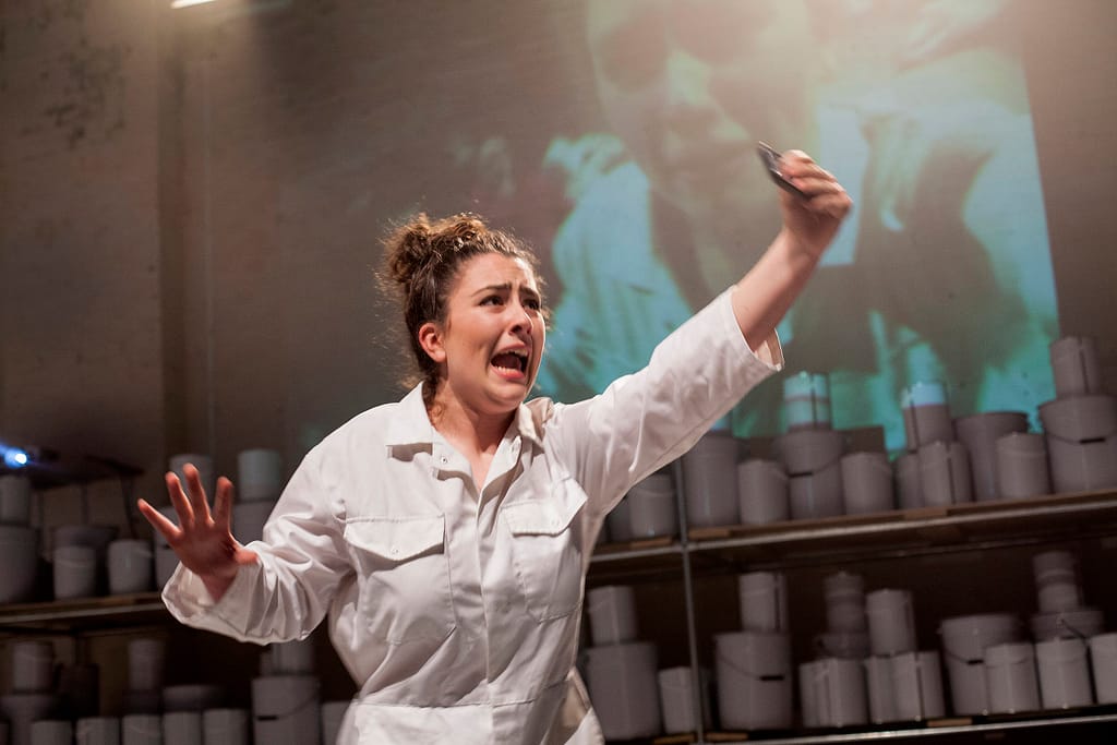 A performer wearing a white boiler suit holds their arm outstretched in front of them, clasping a mobile phone while screaming. There are projected images on the wall behind, with shelving filled with pots/tins