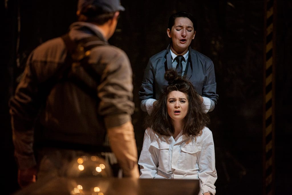 A performer in a white boiler suit sits staring distressingly at the camera. Behind them a person in a smart grey suit and tie runs their hands through their hair.