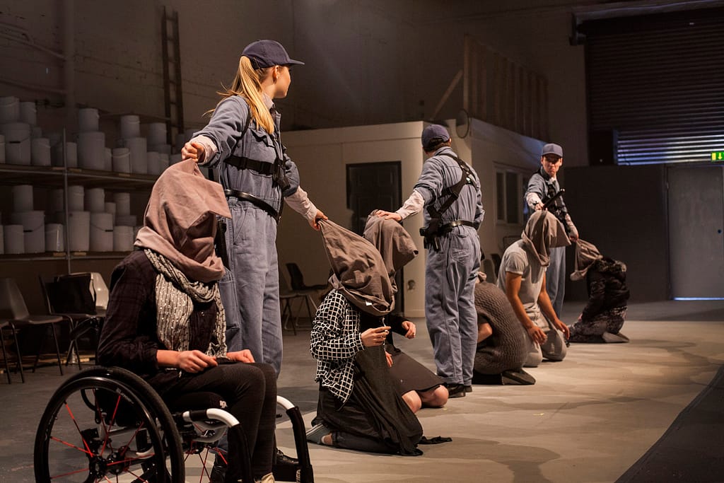 Performers dressed in grey stand in a line. Half have sacks over their heads while kneeling on the ground. The other performers stand there awaiting to remove the restrictive sacks