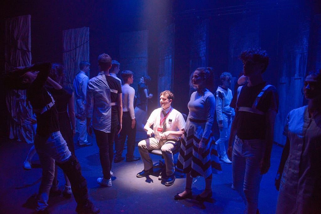 A figure dressed smartly in shirt and tie sits on an office chair in the middle of a stage under a spotlight. They are circled by a crowd of performers menacingly