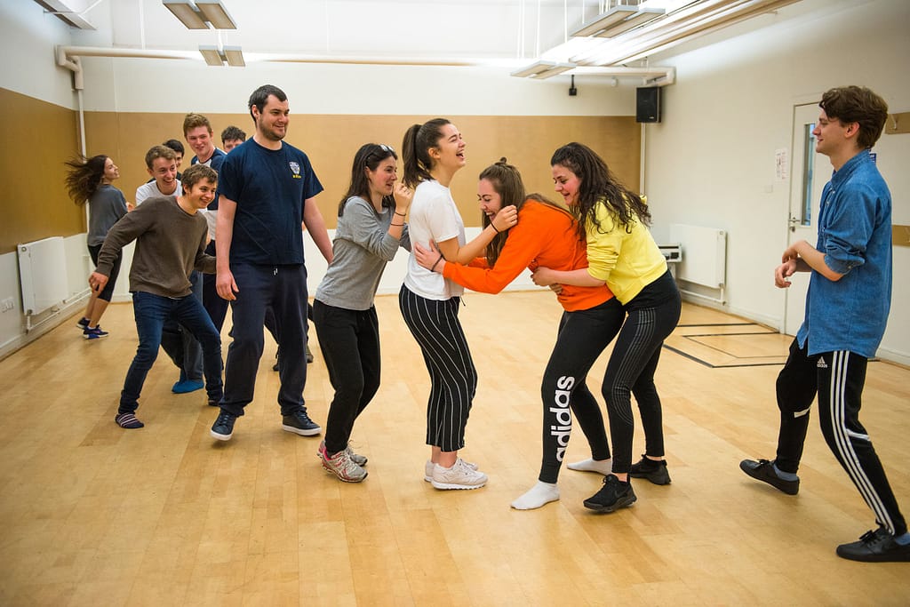 Two conga lines of young people face one another and embrace in the middle, laughing and moving in a brightly lit room with wooden floors