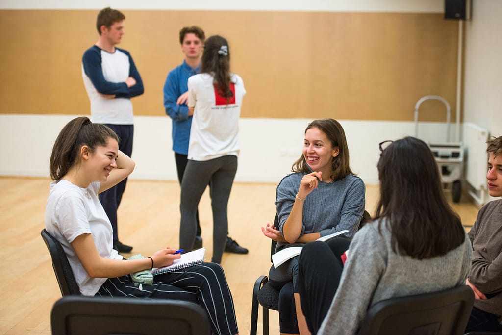Four young people sit on chairs facing one another, with scripts on their lap, smiling and talking. In the background, 3 other figures stand in a circle with arms crossed, talking to one another