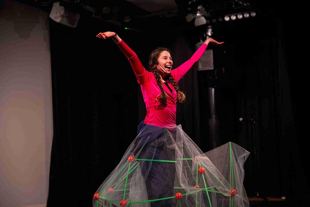 'ENCOUNTERS' – Performer stands on stage with arms stretch looking outwards. They have long dark hair and is worn in plaits. They wear a pink top, dark blue bottoms and a stage microphone. On their lower half is a structural skirt made of green sticks and orange balls with a sheer white netting over it.