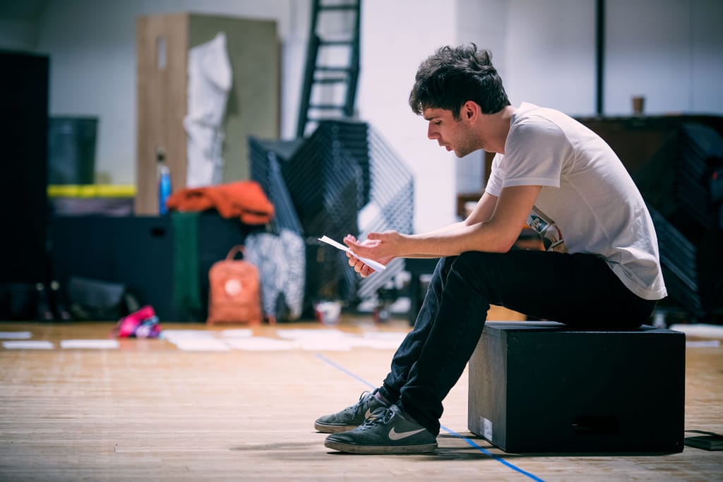 A young performer sits on a black box on a wooden floor, dressed in a white t-shirt and black trousers, holding a sheet of paper rehearsing lines