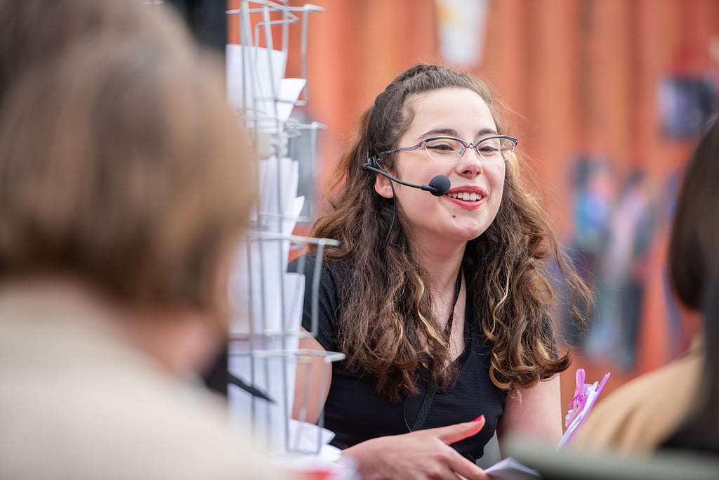 A young person wearing glasses and a head mic stands next to a leaflet stand and smiles at someone while holding a pink clipboard