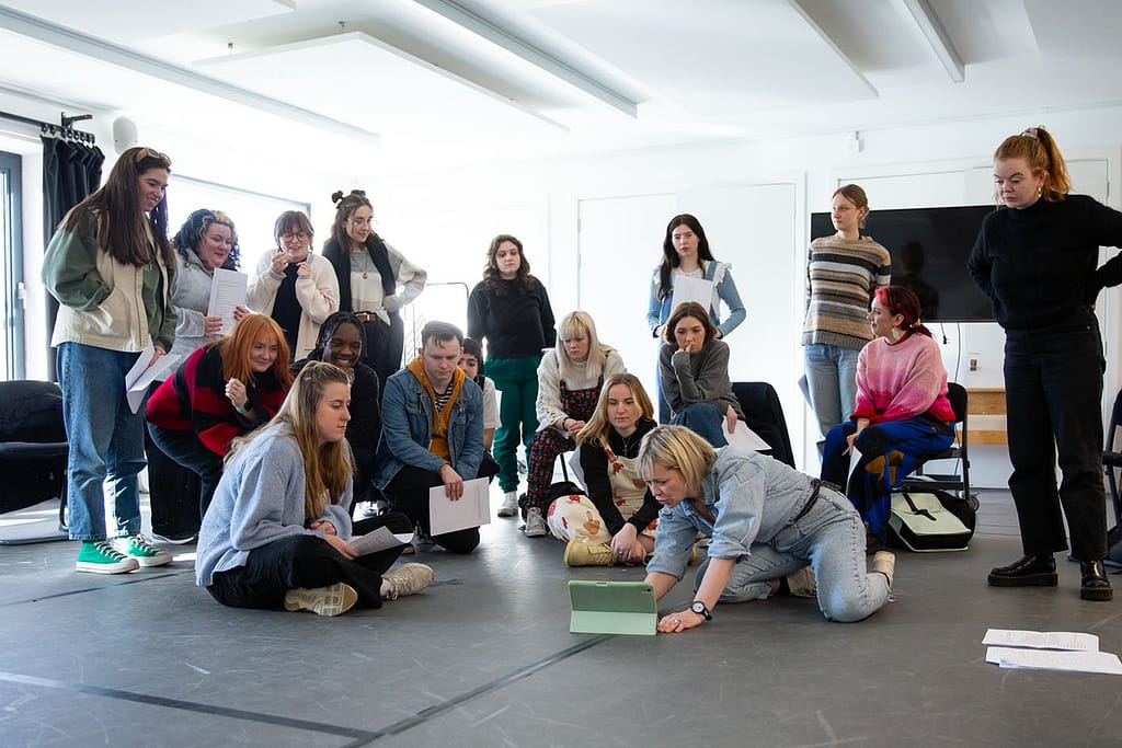 A group of young people crowd around an iPad placed in the middle of the floor between them. Some sit crossed legged on the floor, some sit on chairs, some are standing. A woman with short blonde hair in denim dungarees is kneeling next to the iPad adjusting it.
