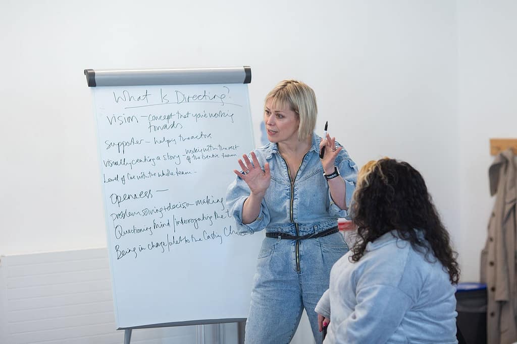 A woman with short blonde hair wearing denim dungarees stands next to a stand of flipchart paper. She has her hands up in front of her with a pen in one hand. There is writing on the flipchart behind her. A young person with long dark hair sits in front of her, looking at her with their back to the camera