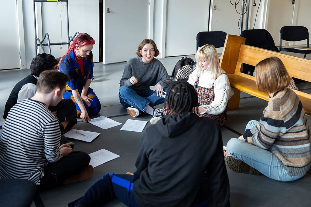 A group of young people sit crossed legged on a grey floor in a circle, with sheets of paper and pens placed in front of them. There is a wooden bench to the right of frame and white doors in the background. The young people are smiling