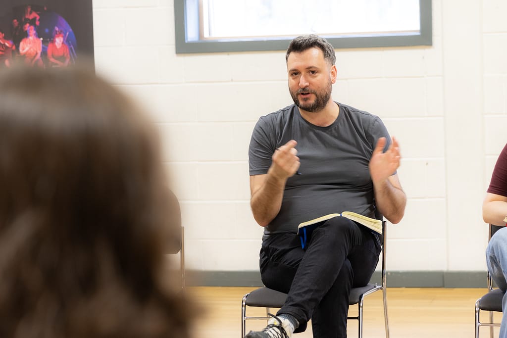 A man with dark hair and a beard sits on a metal chair with his legs crossed and a notebook on his lap. He gestures towards the camera with a pen in his right hand. He is wearing dark jeans and a dark grey t-shirt.