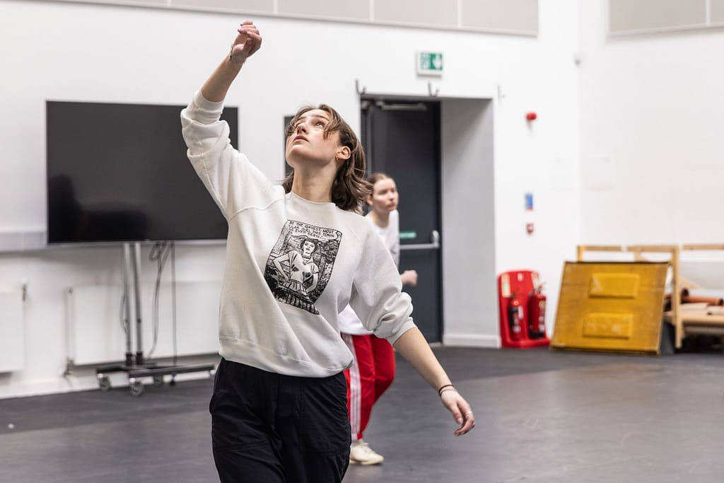 A young person in black trousers and a white sweatshirt with medium-length brown hair looks up towards the sky, gesturing upwards with their right hand. In the background, another person in red track bottoms and a white t-shirt leans forward, mid movement, with their arm crossed over their body.