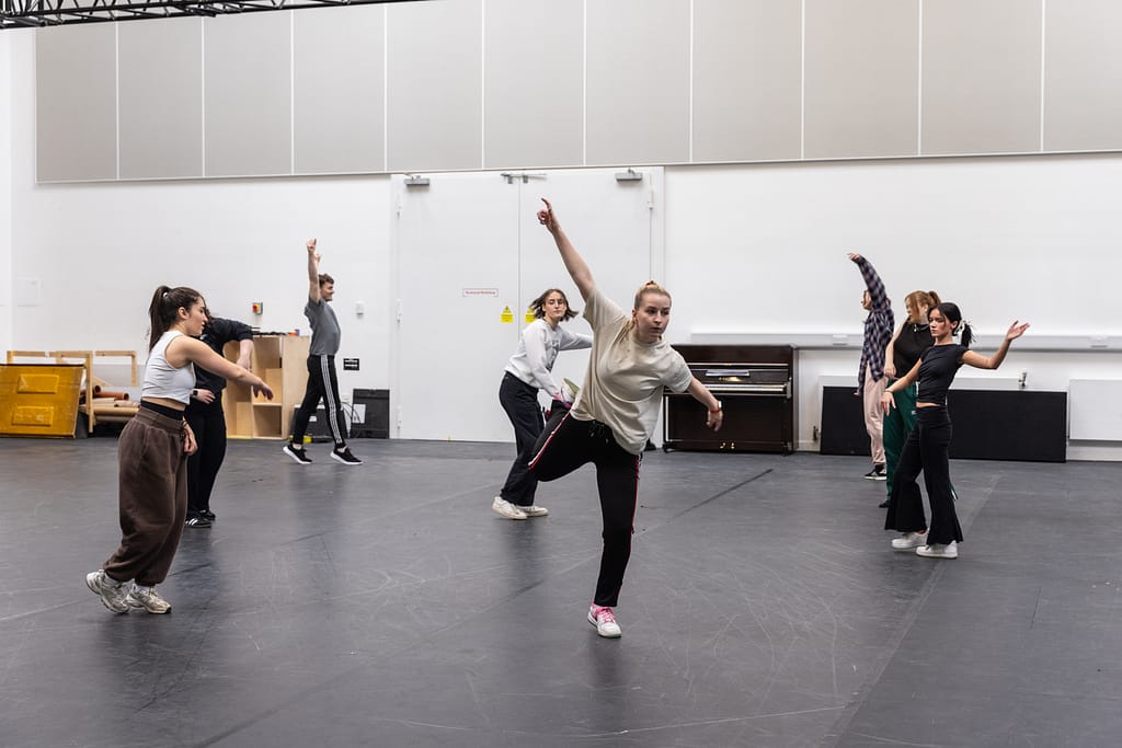 A group of performers stand roughly in a circle facing different directions, all mid-movement. Some stretch their arms towards the ceiling, some out to the side, one spins on the spot while the performer in the centre leans on one leg whilst pointing their hands in opposite directions. They are in a white walled room with black flooring.