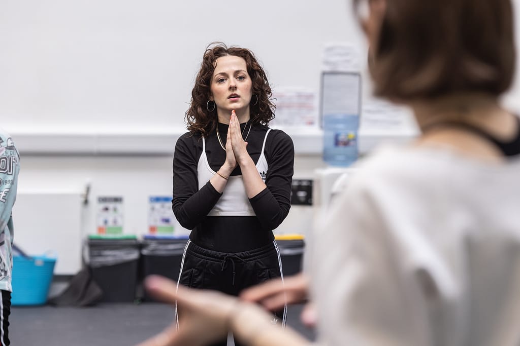 A woman with curly, brunette hair wearing black track bottoms, black long sleeved top and white tank top looks towards the camera inquisitively with her hands clasped in front of her