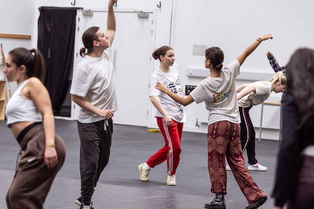 5 performers wearing white t-shirts and various coloured tracksuit bottoms move dynamically around the space, facing in different directions and jiving their bodies using their arms, shoulders, legs and heads.
