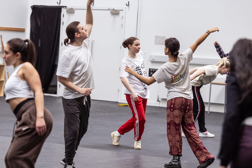 5 performers wearing white t-shirts and various coloured tracksuit bottoms move dynamically around the space, facing in different directions and jiving their bodies using their arms, shoulders, legs and heads.