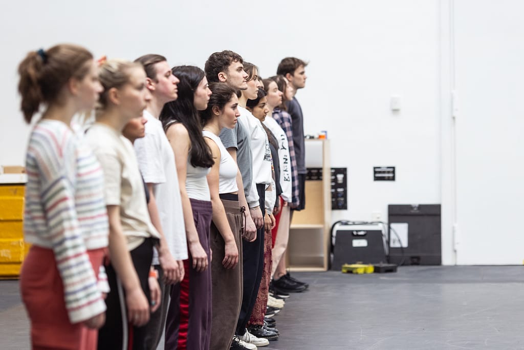 A group of young performers stand to attention in a line to the left of frame, all looking in front of them.