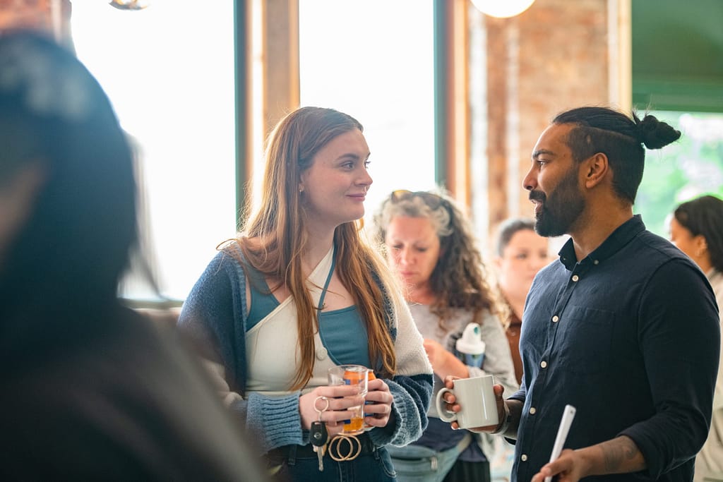 Two audience members are talking to one another. Both are holding the drinks and standing in front of a window.