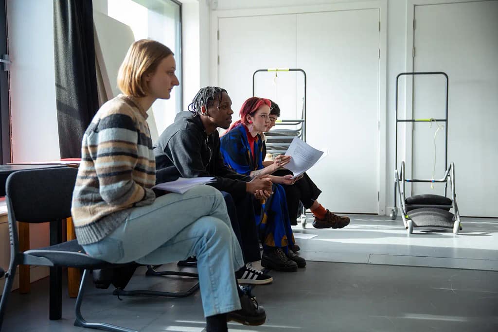 4 young people in a line sit on chairs while leaning forward all facing the same direction. 2 have their legs crossed while 2 hold scripts of paper in front of them.