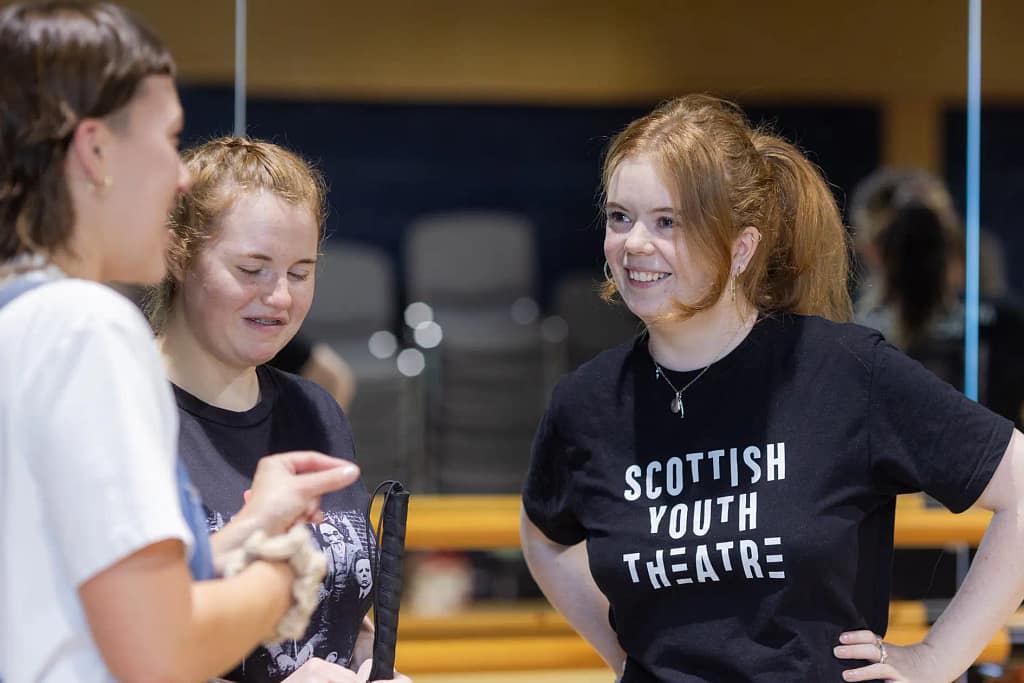 A young woman with red hair wearing a black t-shirt with a Scottish Youth Theatre logo on it smiles towards another person, with her hands resting on her hips. Two other people stand next to her smiling.