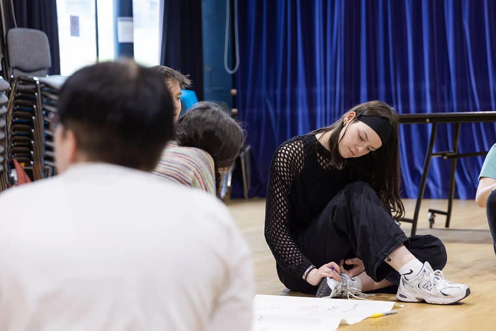 A girl dressed in black sits on a wooden floor with her legs crossed under her. She looks down to her right at a sheet of paper on the floor. Other figures are in the frame, with a blue curtain in the background.