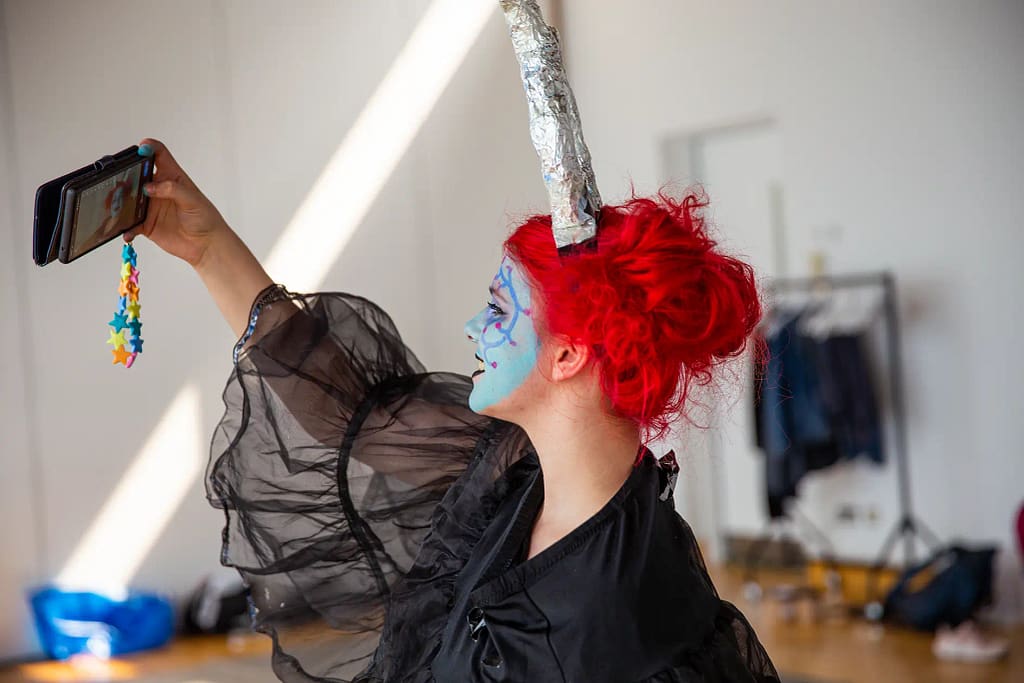 A young person with bright red hair tied up in a bun faces away from the camera, taking a selfie on their phone. They are wearing a black dress with sheer sleeves, horns on their head made from foil and light blue decorational face paint.