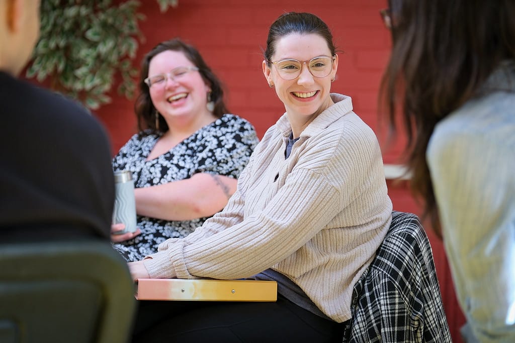A white woman in glasses smiles at someone sitting in front of her whilst sitting at a table. Another person with glasses and long hair laughs.