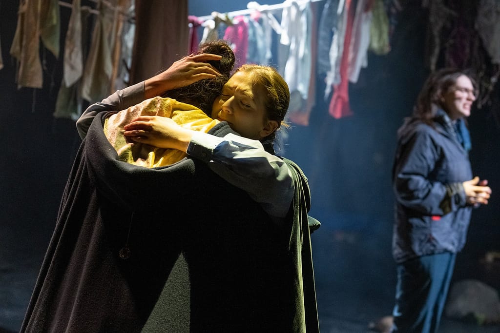 On a stage an actor holds another actor in an embrace, both wearing capes. Another actor stands behind them, addressing the audience.