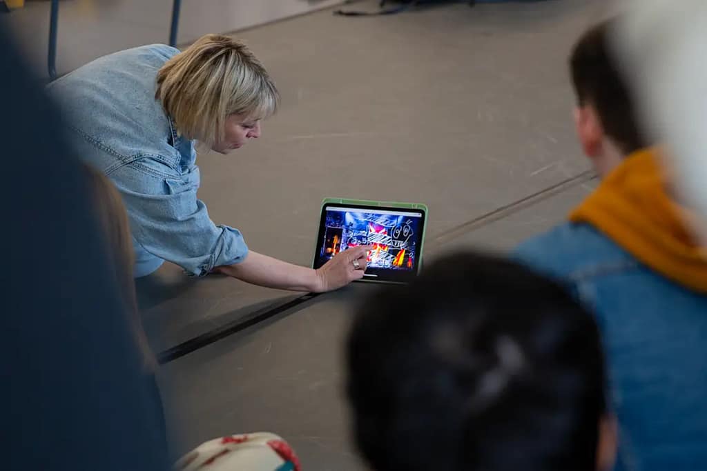 A woman in denim overalls leans over on the floor, touching a tablet which is propped up and resting on the floor. She is touching the screen which displays an interesting theatre diagram. Various young people look towards her and the screen.