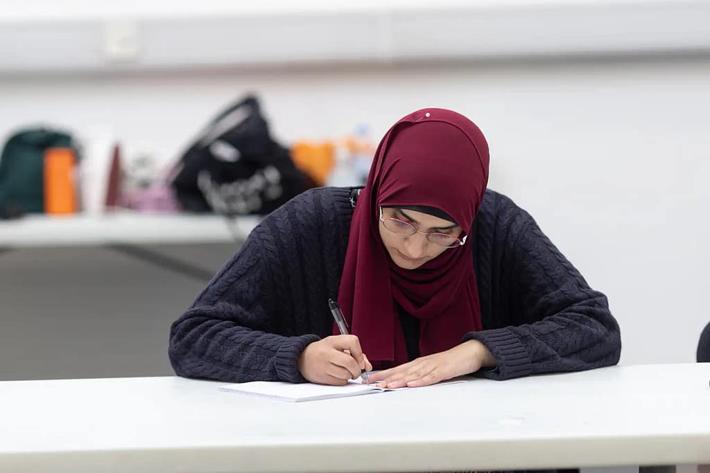 A young woman, dressed in a dark blue knitted jumper and maroon hijab while wearing thin, red framed glasses, sits at a white table and leans on a notebook while writing in it with a pen. There is clutter atop another table in the background.