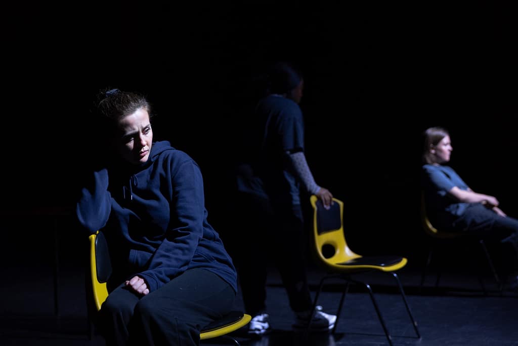 A dark stage with three performers, two sitting on chairs one standing behind it. The lighting has dark shadows on their faces and the performers faces look troubled.
