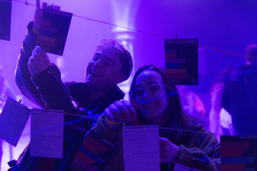 Two audience members bathed in blue light, laugh as they clip small pieces of card to strings that are going in different directions across the frame.
