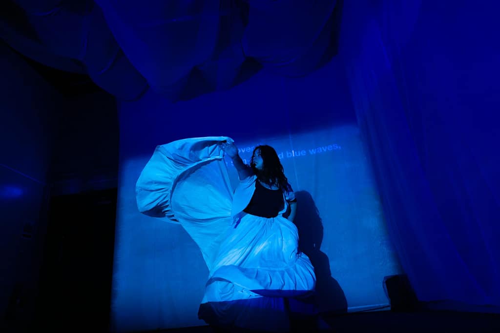A performer dances, bathed in a blue and a projection. They wear a long white, skirt and dance with it above their head.