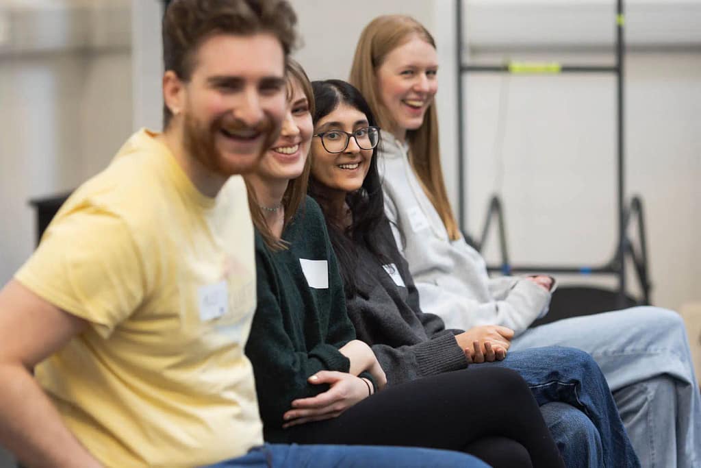 A group of 4 young people wearing jumpers and t-shirts sit in a row next to one another, looking towards the camera smiling and laughing. The all have one leg crossed over the other in front of them, with their hands clasped/folded on their lap