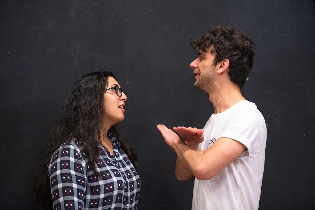Two actors face one another, closer together against a black wall. One performer is gesturing in a way that suggests they are having a disagreement