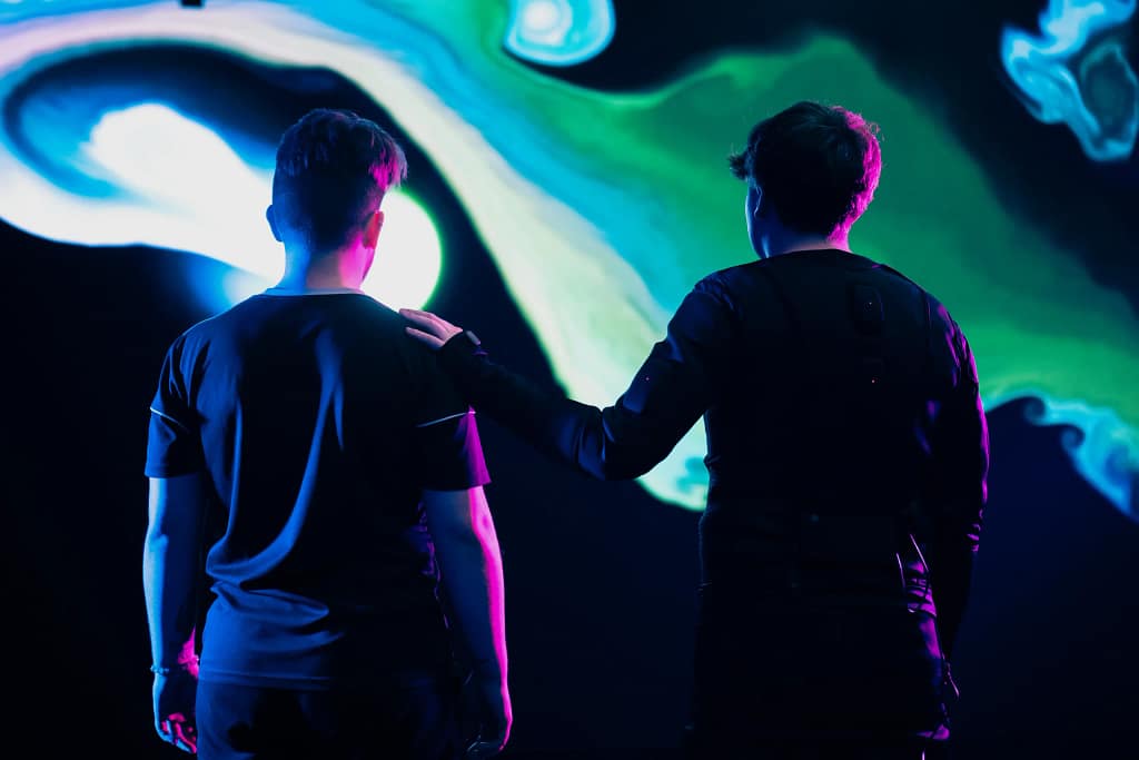 Two performers with their backs to the camera, silhouetted by a neon pink light, stand in front of a large digital screen with organic green, blue and white shapes on it. One performer rests their hand on the shoulder of the other performer.