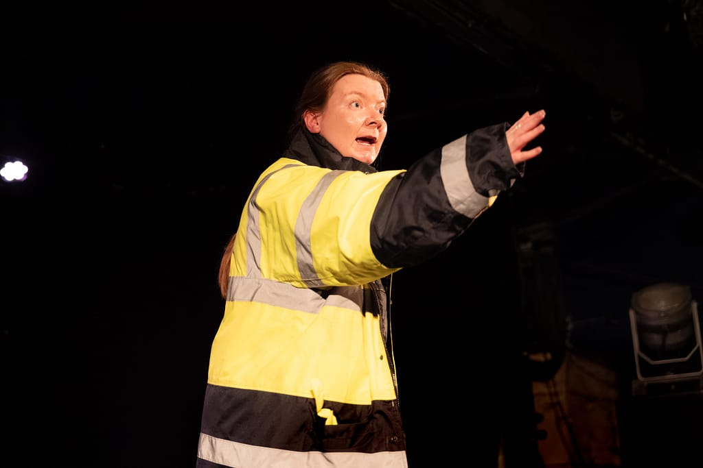 A performer stands on stage, wearing a high-vis jacket, and gestures frantically out to the audience.
