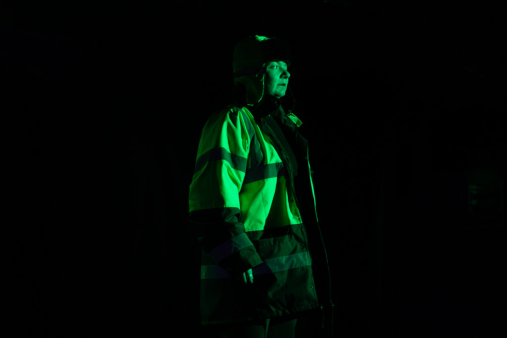 A performer stands on staged, covered in green moody light, wearing a high-vis jacket and hat