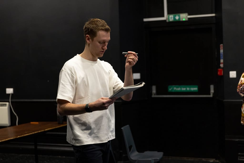 The director stands in a black-box room in white t-shirt and gestures looking down at a script in his hand.