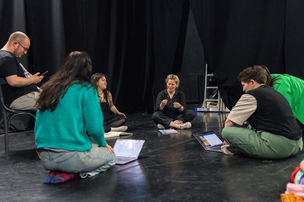 A group of young people sit mostly cross legged on a black stage floor, in a circle, all with laptops, phones or notebooks placed in front of them as they smile at one another