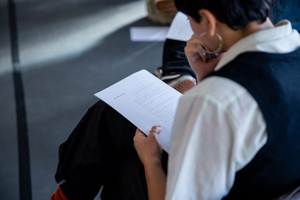 Over the shoulder of a figure with a script on their lap. They have short dark hair, a gold hoop earring in their left ear, a ring on their left thumb and their legs crossed. They are wearing a white collared shirt and black sweater vest.