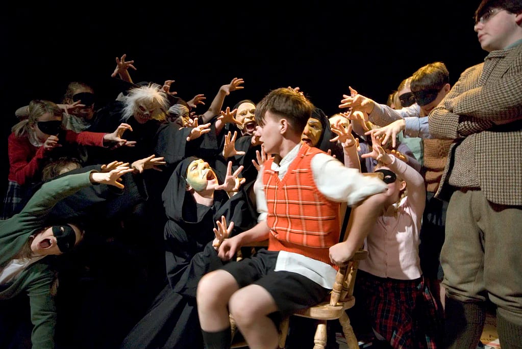 A young boy in dark shorts, a white shirt and tartan sleeveless sweater sits in a wooden chair looking panicked, surrounded by performers in ghoulish masks who leap towards him